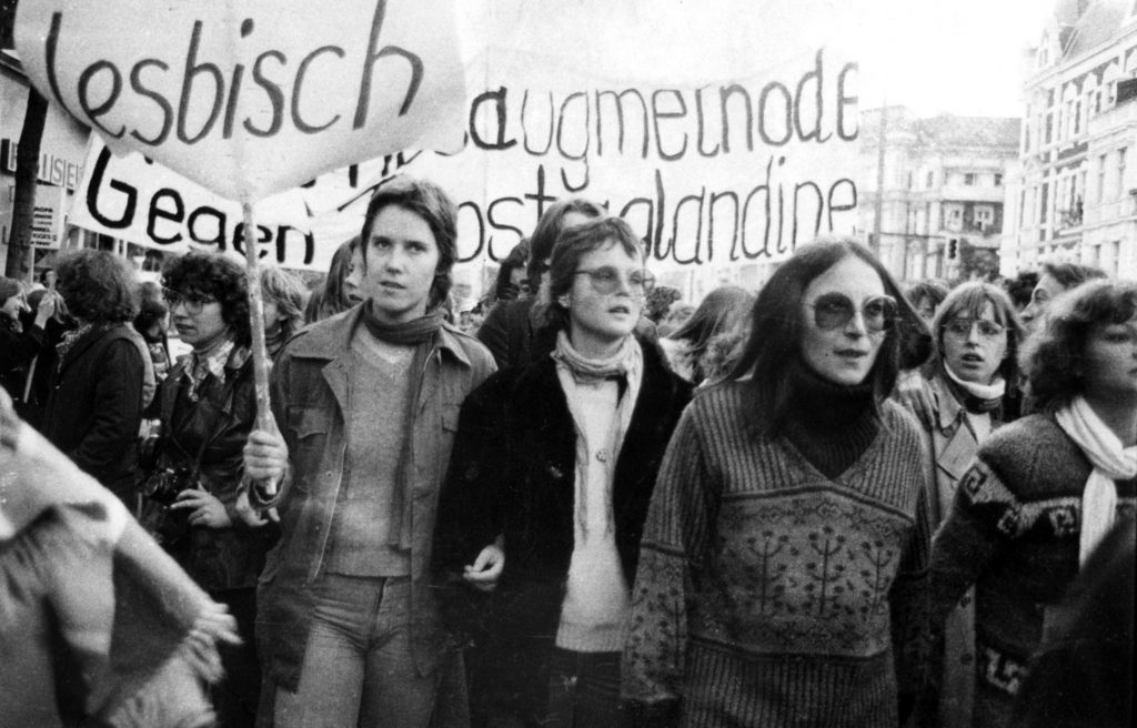 Zu sehen ist eine Schwarz-Weiß Fotografie einer Demonstration gegen den § 218. Im Vordergrund läuft eine Gruppe Frauen, eine von ihnen hält ein Transparent auf dem „Lesbisch“ steht, weitere Transparente sind im Hintergrund. 